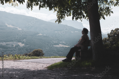An elderly woman sits under a tree and enjoys the Alps panorama. Sunny view with some hazy sky