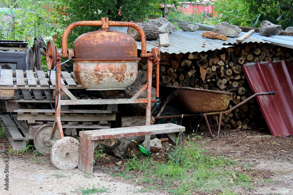 Electrical metal concrete mixer covered with rust next to completely rusted wheelbarrow construction cart surrounded with construction material and firewood in backyard on warm sunny day Electrical metal concrete mixer covered with rust next to completely rusted wheelbarrow construction cart surrounded with construction material and firewood in backyard on warm sunny day