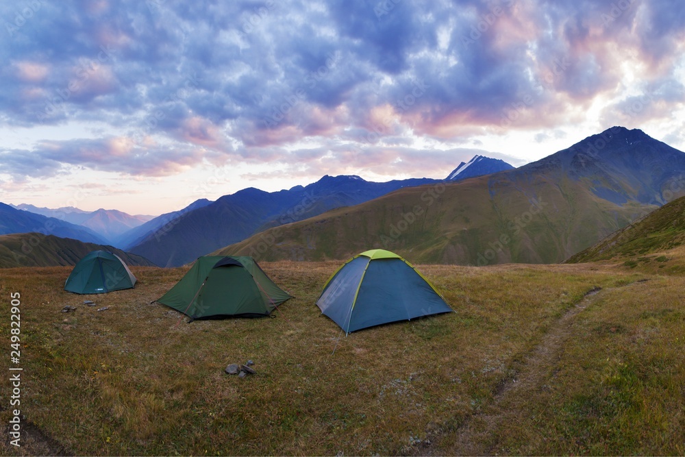 Colorful summer panorama with green tourist Tents on the hill beneath the mountains under blue sky. Beautiful mountain wild concept