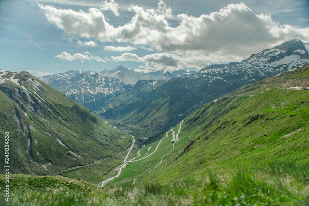 Sunny panoramic picture above Andermatt in Switzerland. A few streets towards the valley, green meadows and a few clouds.