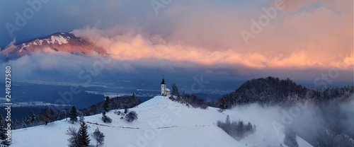 Panoramic view of beautiful winter wonderland mountain scenery in the Alps with pilgrimage church of Jamnik and famous Julian Alps in the background  Bled  Slovenia. Christmas time in Europe concept