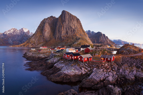Winter landscape with houses in village, city illumination, snowy mountains, sea, blue cloudy sky reflected in water at dusk. Beautiful Hamnoy, Lofoten islands, Norway. Norwegian fishing village