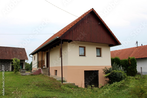 Renovated small suburban family house with new facade and roof tiles partially made of wood with large basement surrounded with grass and other structures in backyard on warm sunny day