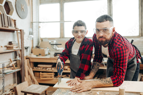 Dad and his son dressed in checkered red shirts and wearing safety glasses are working in the carpentry workshop, son is learning to work with a saw under father s supervision.