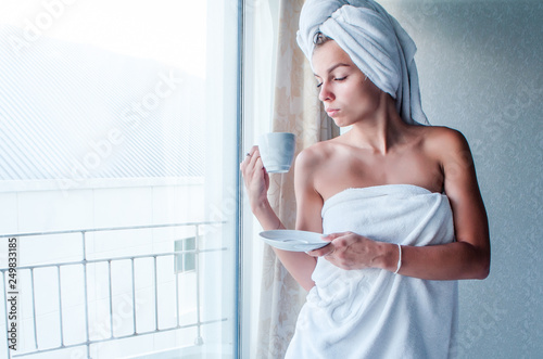 Young caucasian cute smiling woman wearing white bathrobe and turban drinking from cup holding plate in hand in hotel