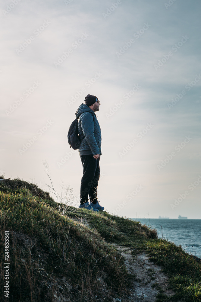 Man traveller with backpack stands on top of rock cliff in grass and looking at sea landscape, freedom of adventure concept