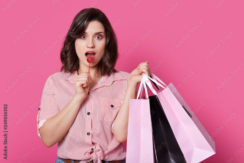 Isolated shot of pinup girl with wavy hair, has surprised expression, enjoys sweet lollipop, holds bags, makes shopping, dressed in stylish shirt, isolated over pink background. Coquettish woman