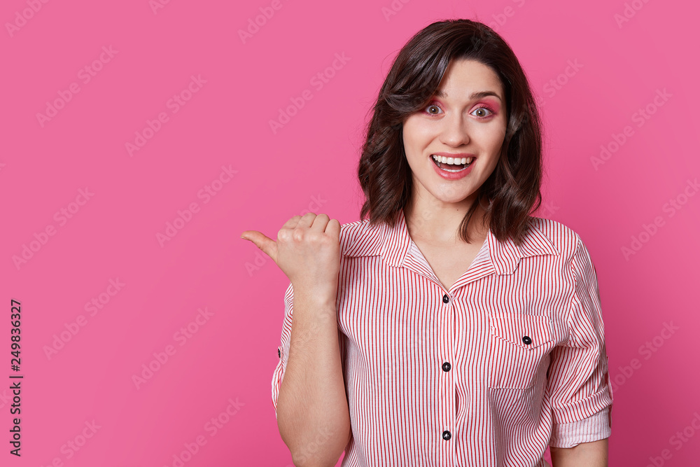 Positive brunette lady with wavy hair, dressed in striped stylish shirt, has make up, points with thumb at blank space, isolated over pink background, promots item or product. People and fashion Positive brunette lady with wavy hair, dressed in striped stylish shirt, has make up, points with thumb at blank space, isolated over pink background, promots item or product. People and fashion