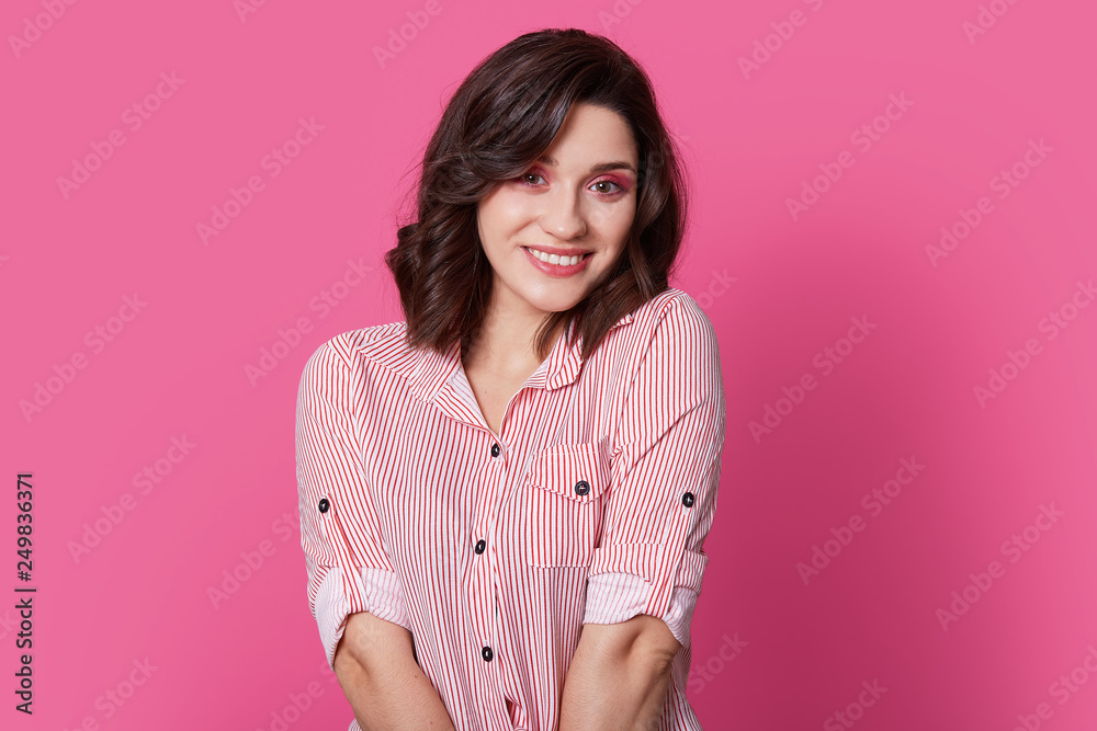 Fototapeta premium Indoor shot of pleasant looking fashionable woman with stylish haircut, makeup, smiles gently, looks with happiness at camera, wears shirt, isolated over pink background. Feminity and style concept