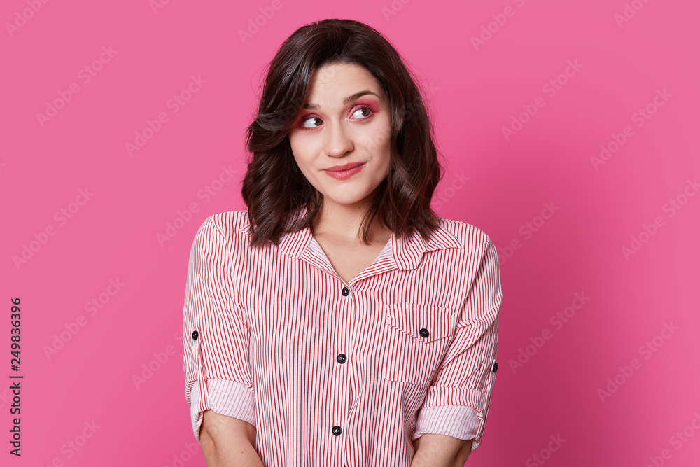 Fototapeta premium Dreamy pleased lady with short hair, looks aside with thoughtful expression, contemplates about something, dressed in fashionable attire, models over pink background. Retro woman models indoor