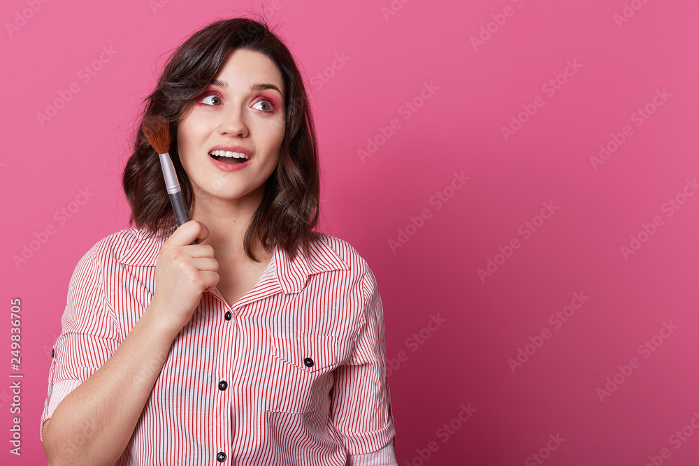 Attractive female model keeps gaze aside, has pink makeup, curly hair, holds cosmetic brush, works in beauty salon, wears striped shirt, isolated over rosy background with blank space aside. Attractive female model keeps gaze aside, has pink makeup, curly hair, holds cosmetic brush, works in beauty salon, wears striped shirt, isolated over rosy background with blank space aside.