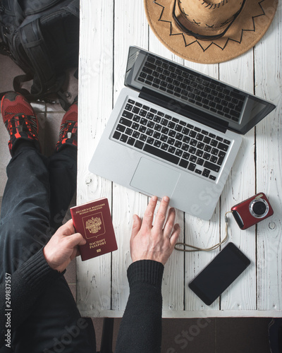 Vacation planning on the computer. A man holds a passport of the Russian Federation. On the white wooden table telephone, camera, hat and glasses