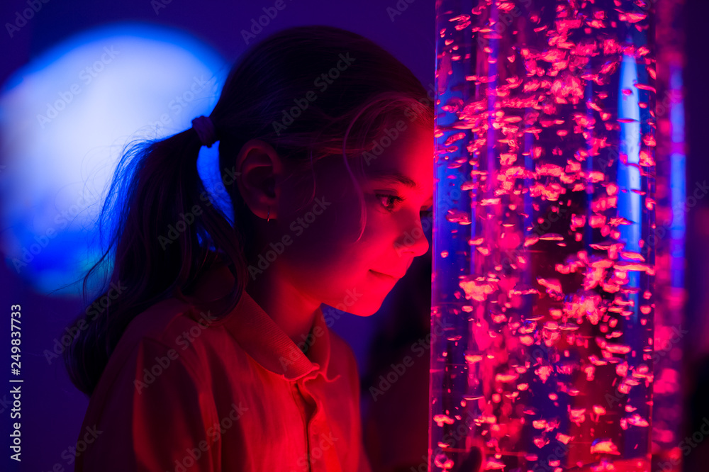 Child in therapy sensory stimulating room, snoezelen. Child interacting with colored lights bubble tube lamp during therapy session. - obrazy, fototapety, plakaty 
