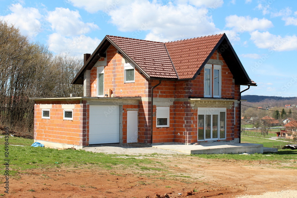 Unfinished under construction small suburban red brick family house with attached garage surrounded with grass and dirt in front with tall trees and houses in background on warm cloudy day Unfinished under construction small suburban red brick family house with attached garage surrounded with grass and dirt in front with tall trees and houses in background on warm cloudy day