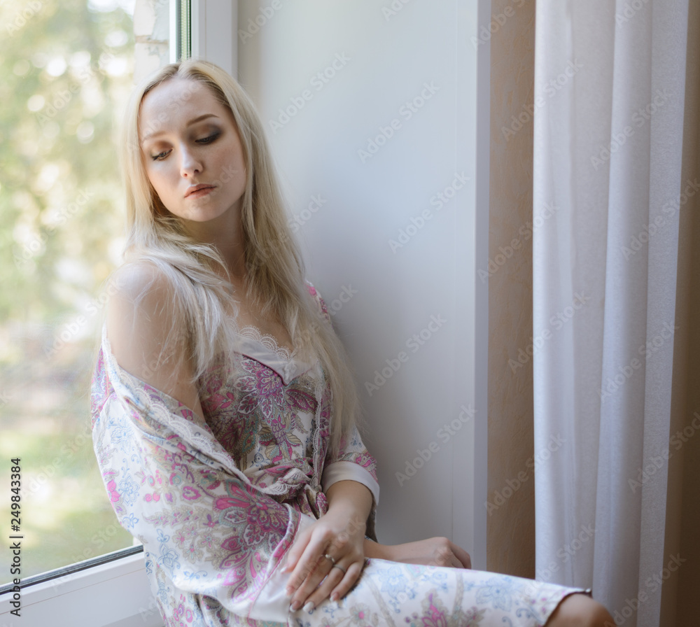 Pensive girl in bathrobe sitting on the windowsill. Natural light. Pensive girl in bathrobe sitting on the windowsill. Natural light.