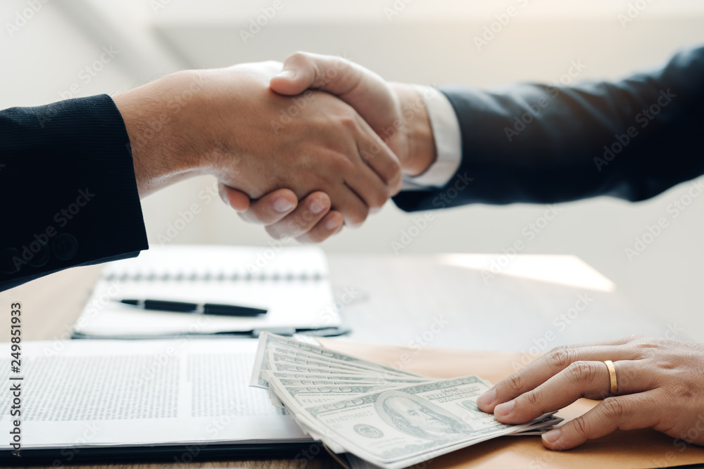 Two corporate businessmen shaking hands while one man places money on document in office room with corruption concept.