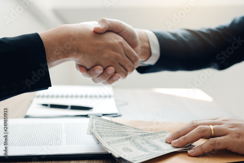 Two corporate businessmen shaking hands while one man places money on document in office room with corruption concept.