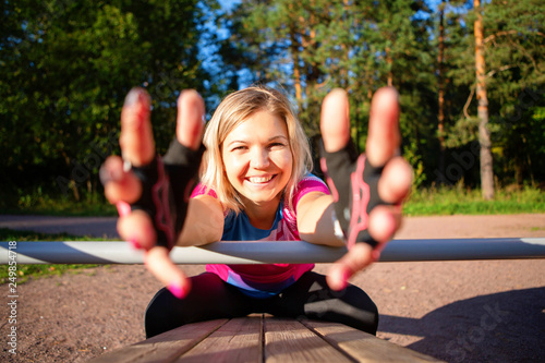 Athlete girl pulling hands forward is exercising at wooden bench in park on summer.