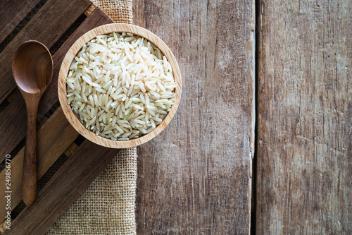 raw brown whole grain semi-milled rice in wood bowl with wood spoon on gunny sack cloth on wooden table, top view with copy space