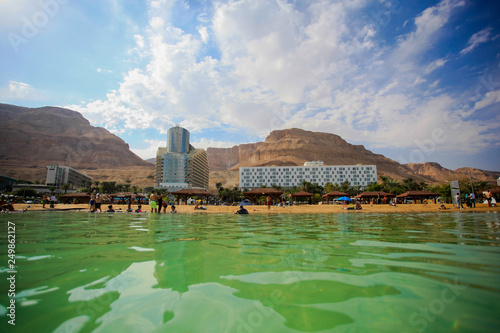 View from the Dead Sea to hotels and beach area. People walk along the beach. Against the background of blue sky and clouds with hills