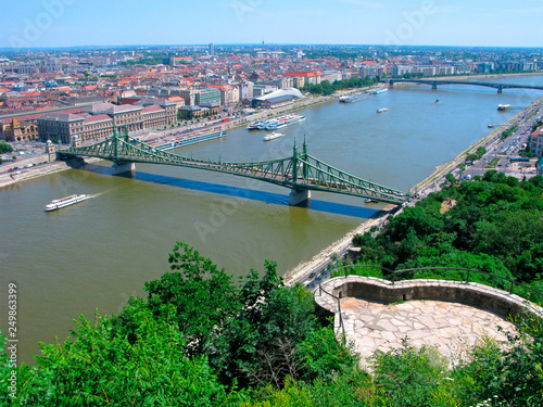 Budapest, Hungary. Panorama of Budapest city with Danube river and Liberty bridge from the Gellert Hill. Observation deck on green Gellert hill. Ships on the river, houses with red roofs.