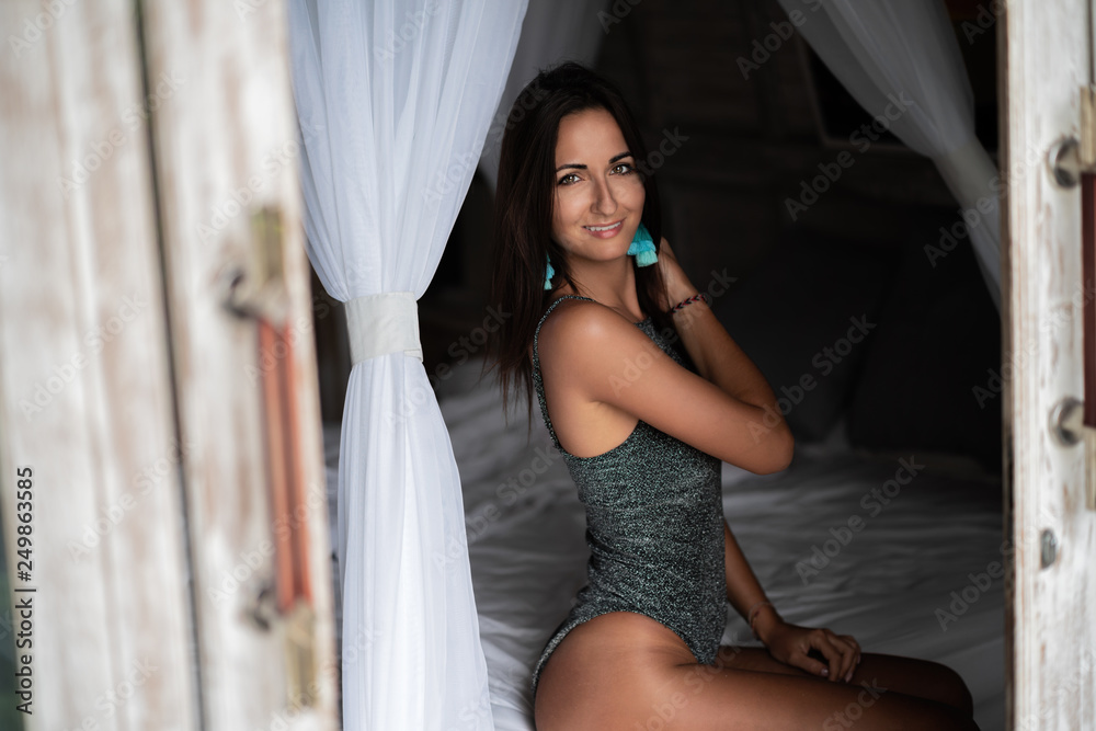 Beautiful tanned girl posing on white bed. The door to the room is ajar. Cool colors Beautiful tanned girl posing on white bed. The door to the room is ajar. Cool colors