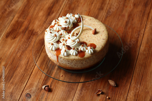 Almond Cheesecake decorated with whipped cream and salted caramel, on round transparent cake stand, on wooden table.
