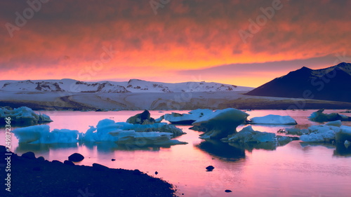 Floating icebergs on Jokulsarlon glacial lagoon with black sand beach in foreground and glacier and red sky during sunset in the background  Southern Iceland.