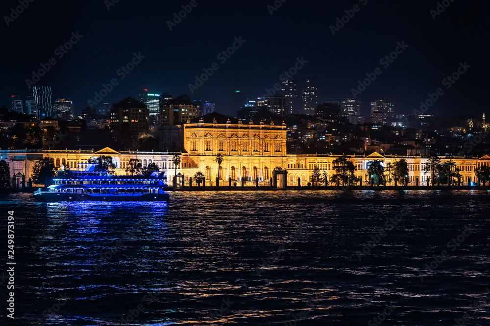 View of the Dolmabahche palace in beams of night-time lighting. Skyscrapers of modern Istanbul at a background and sea ferry on front side is visible.
