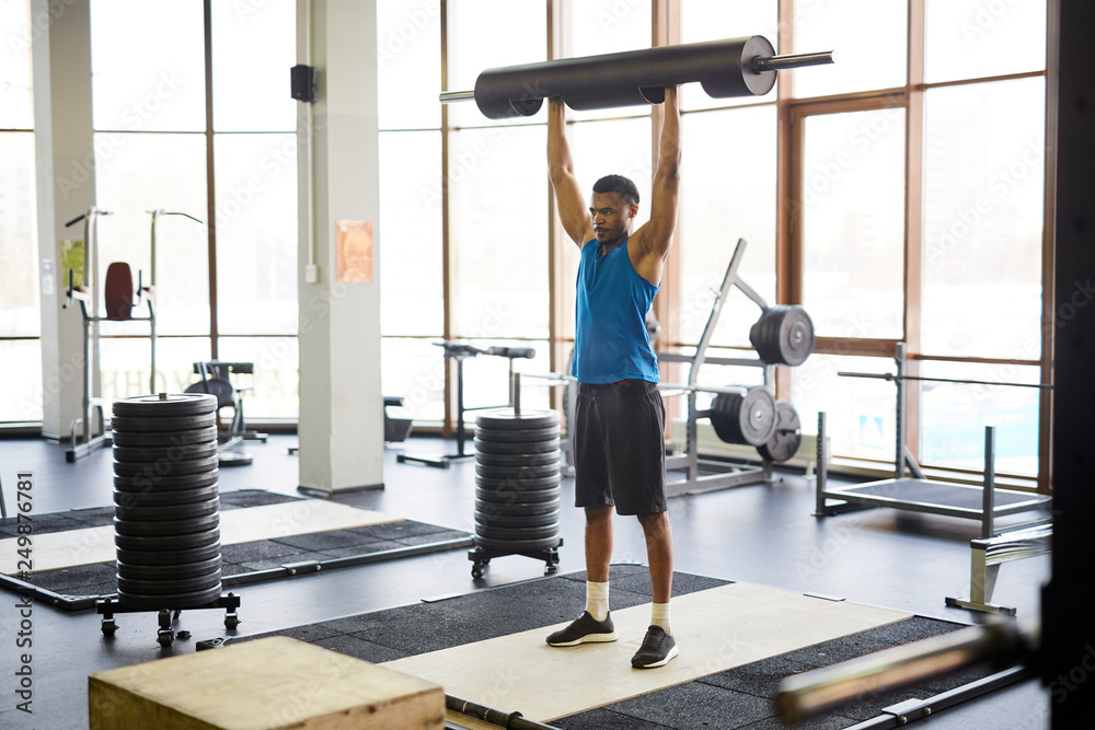 Fototapeta premium Young sportsman in activewear lifting heavy barbell over his head while exercising in cross training center