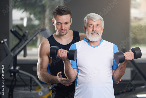 Elderly man and trainer posing with dumbbells in gym.