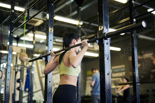 Back view of active brunette girl trying to lift heavy barbell while training in fitness center