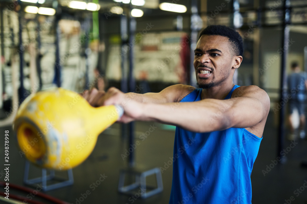Young strong athlete making effort while exercising with heavy kettlebell in fitness center or gym Young strong athlete making effort while exercising with heavy kettlebell in fitness center or gym