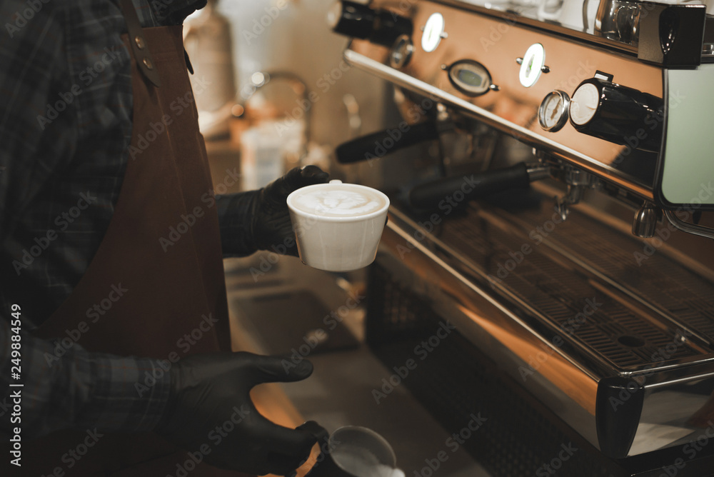 Close up photo of barista in the apron and gloves holding a cup with latte with beautiful latte art. Background photo of bartender at coffee machine with beautiful coffee art. Close up photo of barista in the apron and gloves holding a cup with latte with beautiful latte art. Background photo of bartender at coffee machine with beautiful coffee art.
