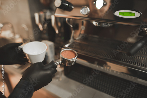 Barista holds in his hand a portafilter with coffee and a cup of coffee, close. Barista Cooks coffee at a professional coffee machine. Preparation of coffee.