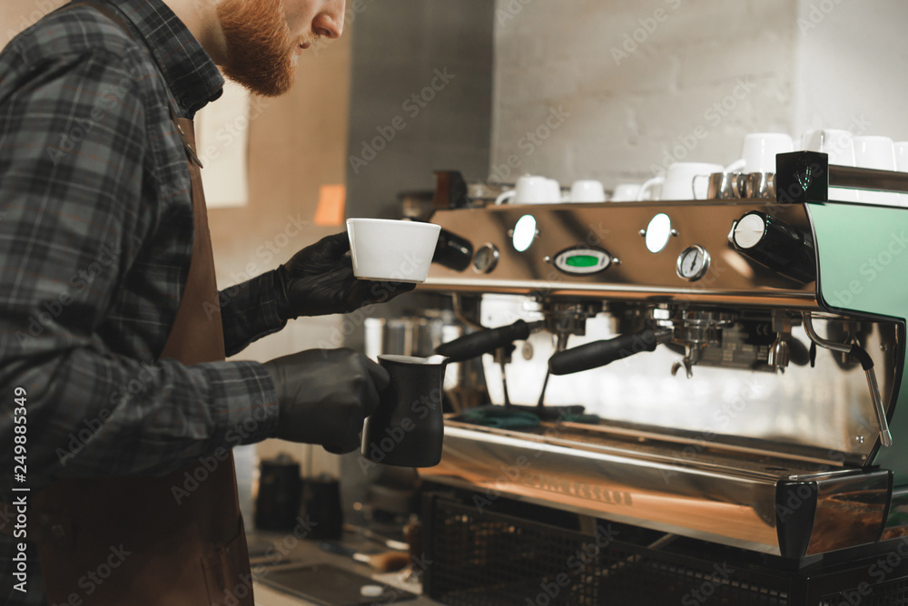 Bearded barista is making a cappuccino at the coffee machine. Close up photo of cafe owner preparing coffee drink. Coffee concept. Bearded barista is making a cappuccino at the coffee machine. Close up photo of cafe owner preparing coffee drink. Coffee concept.