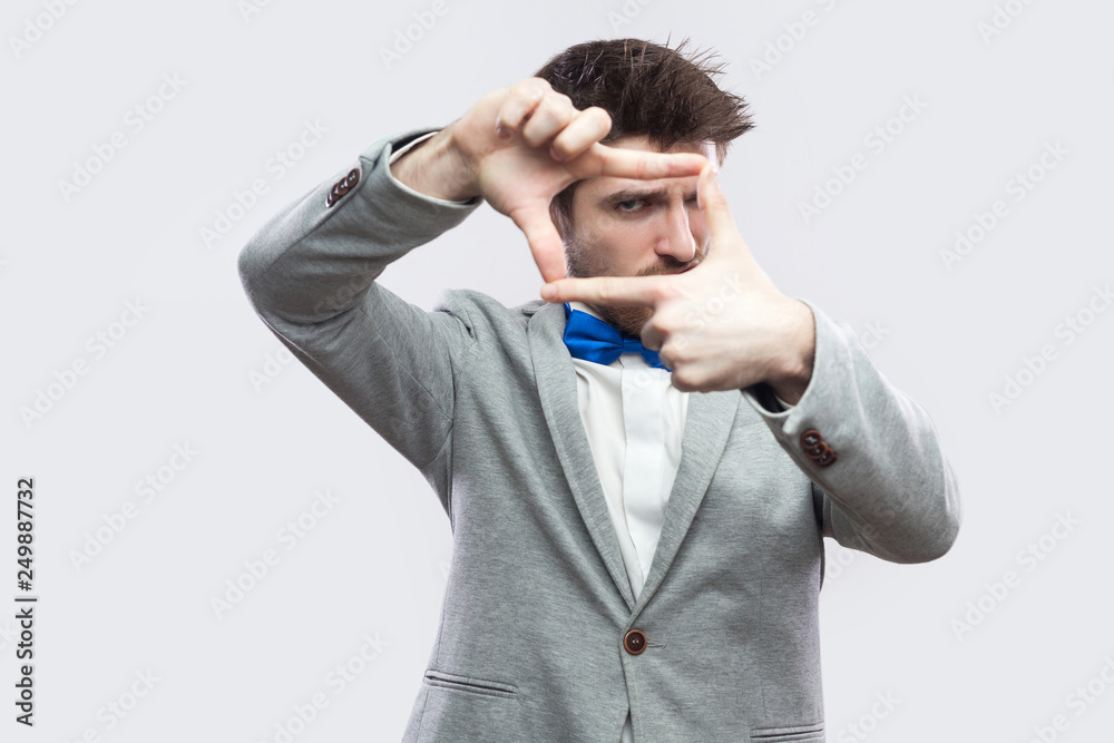 Portrait of serious handsome bearded man in casual grey suit and blue bow tie standing and looking at camera with cropping composition gesture. indoor studio shot, isolated on light grey background.