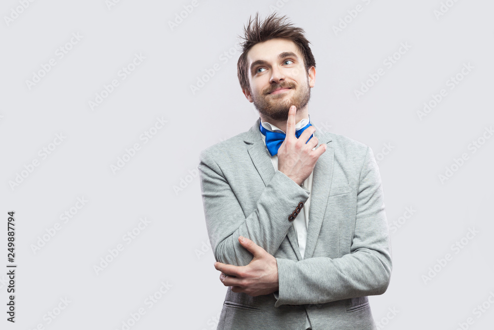 Portrait of dreamful happy handsome bearded man in casual grey suit and blue bow tie standing and looking away, smiling and dreaming. indoor studio shot, isolated on light grey background.