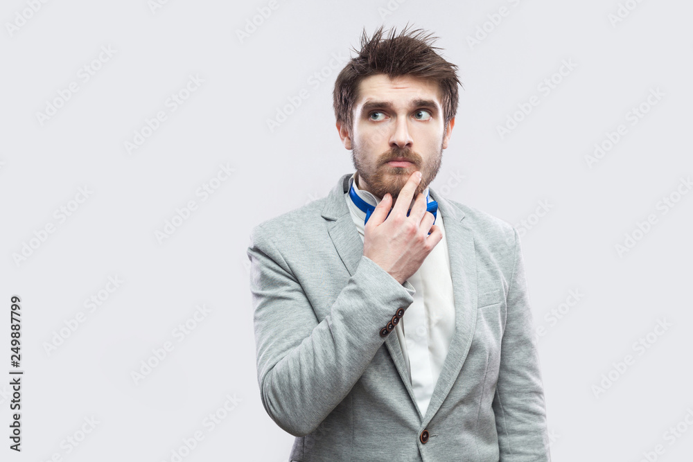Portrait of doubtful handsome bearded man in casual grey suit and blue bow tie standing touching his chin and looking away, thinking. indoor studio shot, isolated on light grey background. Portrait of doubtful handsome bearded man in casual grey suit and blue bow tie standing touching his chin and looking away, thinking. indoor studio shot, isolated on light grey background.
