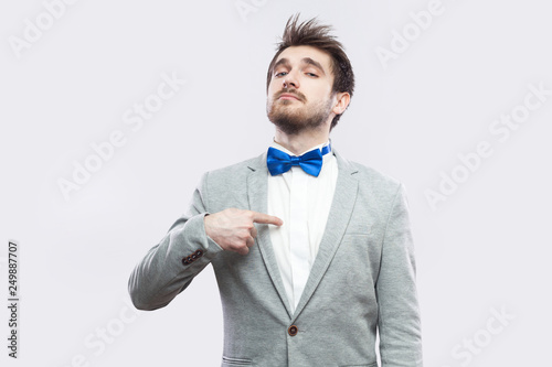 Portrait of proud satisfied haughty handsome bearded man in casual grey suit and blue bow tie standing, pointing himself and looking at camera. indoor studio shot, isolated on light grey background.