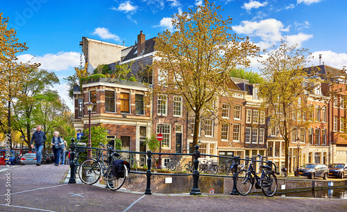 Street of Amsterdam city. Netherlands. Bridge over channel with traditional dutch houses and bicycles. Evening time warm sunlight, blue sky with clouds. Spring cityscape, green and yellow autumn.