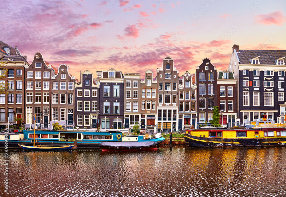 Fototapeta premium Amsterdam, Holandia. Pływające domy i łodzie mieszkalne oraz łodzie na kanałach przy brzegach. Tradycyjne holenderskie tańczące domy wśród drzew. Wieczorem jesień ulica nad wodą różowe niebo zachód słońca z chmurami.