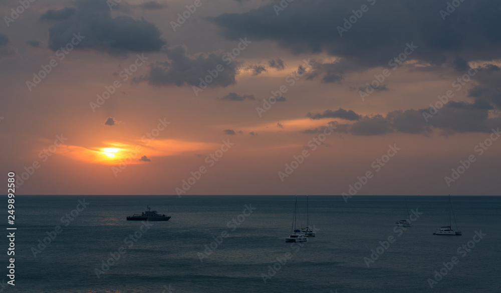 A group of boats is anchoring at the coastline of Koh Lanta, Thailand. The sun goes down in the background.