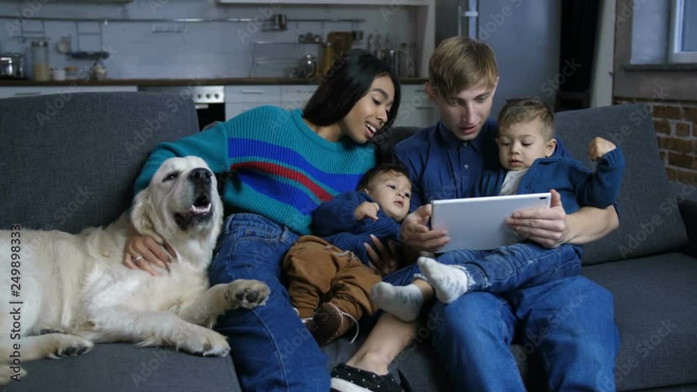 Positive multinational family with adorable toddler sons in hug with labrador retriever dog streaming cartoons online using digital tablet at home. Lovely diverse family and pet dog relaxing on sofa. Positive multinational family with adorable toddler sons in hug with labrador retriever dog streaming cartoons online using digital tablet at home. Lovely diverse family and pet dog relaxing on sofa.