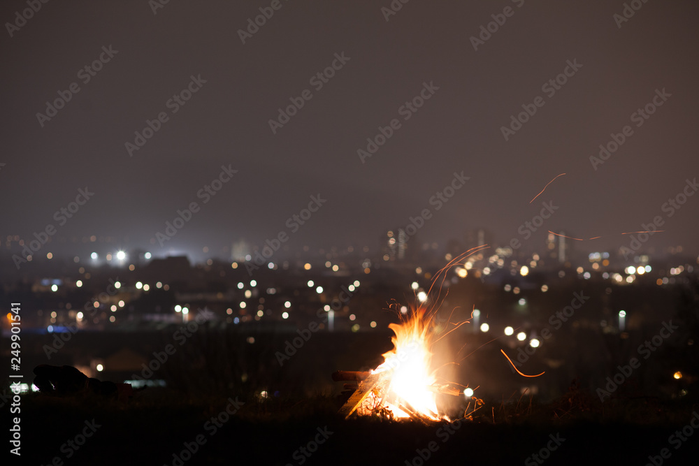 A glowing campfire on top of a hill, with the city lights below
