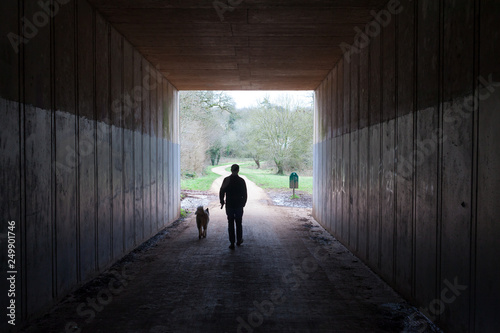 A man and his dog walking out of a tunnel into a bright green park