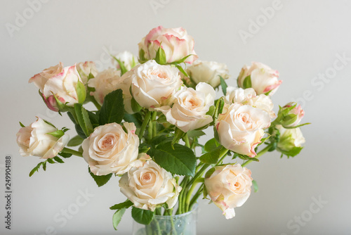 Close up of pale pink roses with green leaves in glass vase against neutral wall background (selective focus)