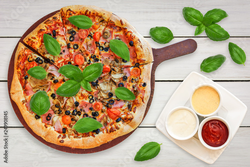 Top view of a traditional italian pizza with: ham, mushrooms, cherry tomatoes, mozzarella, black olives decorated with basil leaves and set of sauces.