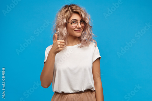 Picture of good looking friendly young Caucasian female wearing stylish clothes and round eyeglasses making approval gesture, showing thumbs up sign at camera and smiling happily, posing at blue wall