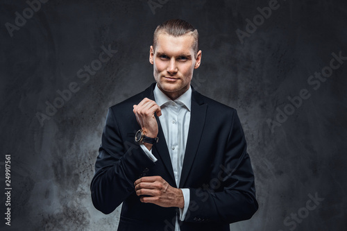 Elegantly dressed handsome man wearing a black classical suit, fastening buttons on the sleeves in dark studio.
